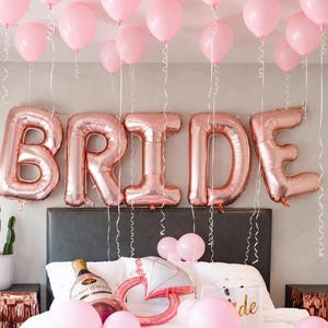 May include: A collection of light pink latex balloons floating above large rose gold foil balloons spelling the word BRIDE. On the bed, there are additional pink balloons, a champagne bottle-shaped balloon, and a diamond ring-shaped balloon.