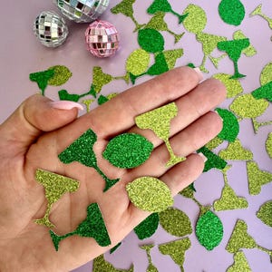 May include: A hand holding green and yellow glitter confetti shaped like limes and margarita glasses. The confetti is scattered on a purple surface.