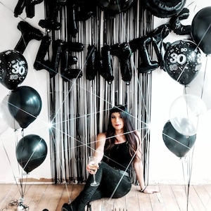 May include: A woman in a black tank top and jeans sits in front of a black and white backdrop with black balloons that spell out "Happy 30th".