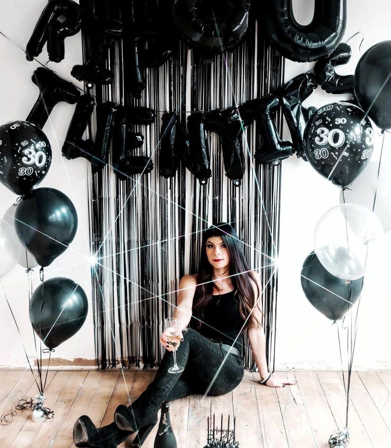 May include: A woman in a black tank top and jeans sits in front of a black and white backdrop with black balloons that spell out "Happy 30th".