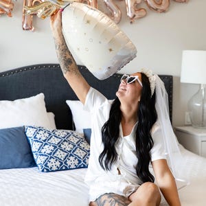 May include: A large champagne bottle-shaped balloon with gold accents and star details is held up. The word "PROSECCO" is spelled out in rose gold letter balloons in the background. The scene suggests a celebration.