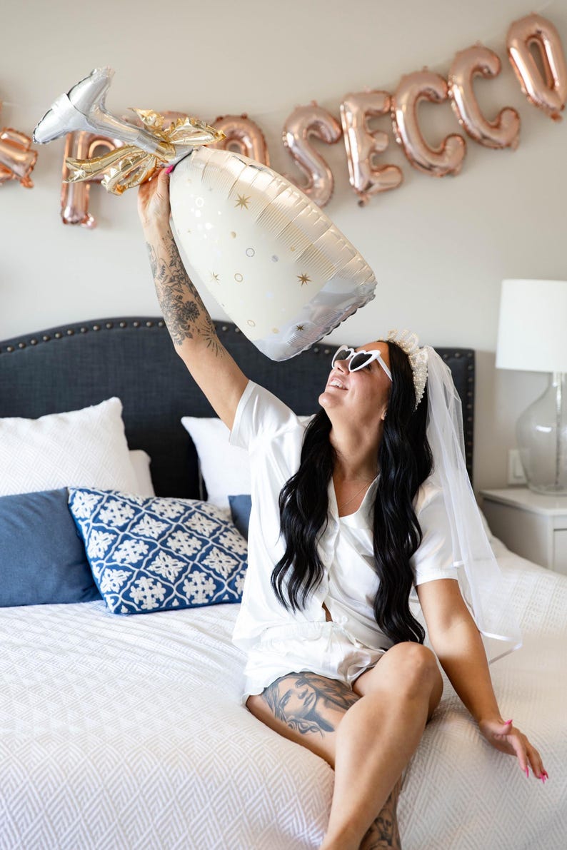 May include: A large champagne bottle-shaped balloon with gold accents and star details is held up. The word "PROSECCO" is spelled out in rose gold letter balloons in the background. The scene suggests a celebration.