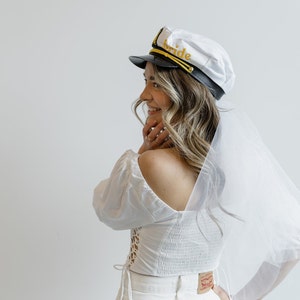 Puede incluir: Un velo de novia blanco con un gorro de capit&aacute;n blanco que dice "bride" en letras doradas. El gorro tiene una visera negra y ribetes dorados.