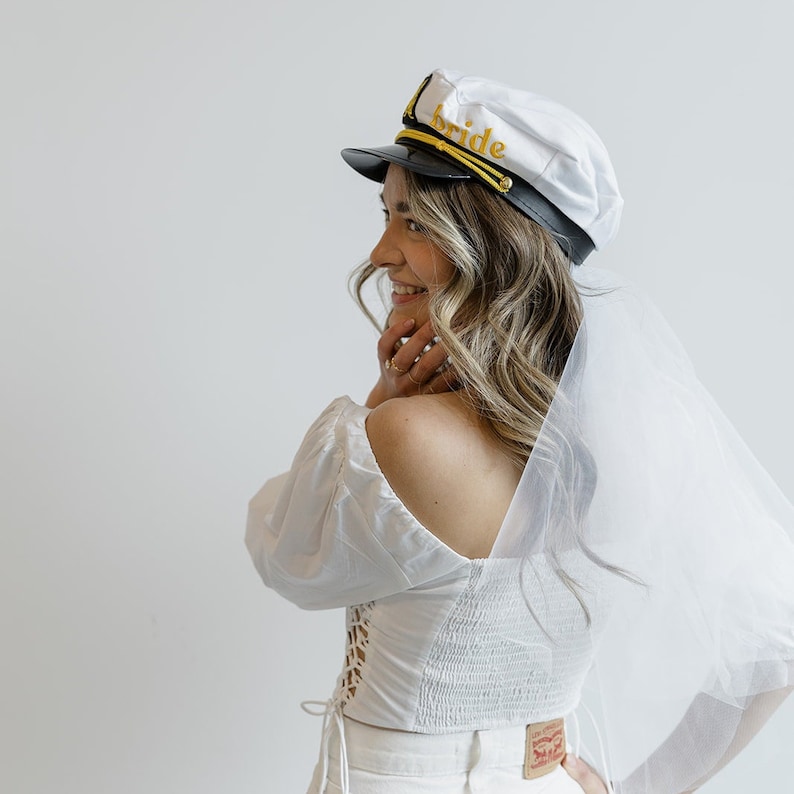 Puede incluir: Un velo de novia blanco con un gorro de capit&aacute;n blanco que dice "bride" en letras doradas. El gorro tiene una visera negra y ribetes dorados.