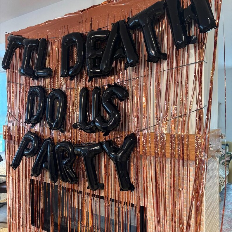 May include: A set of black foil letter balloons spelling out the phrase 'TIL DEATH DO US PARTY' arranged in three rows. The balloons are mounted against a backdrop of shimmering metallic rose gold fringe curtains that hang in vertical strips.