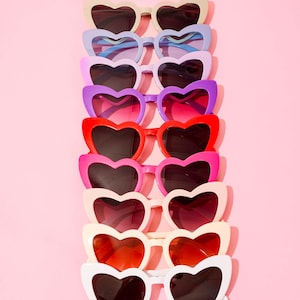 May include: A collection of heart-shaped sunglasses in various colors, including white, red, pink, purple, and blue, arranged vertically against a pink background. The sunglasses have dark lenses and a playful, fashionable aesthetic.