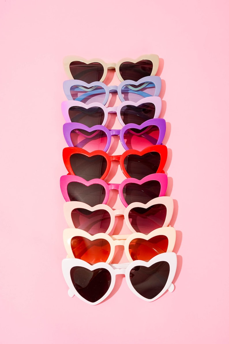 May include: A collection of heart-shaped sunglasses in various colors, including white, red, pink, purple, and blue, arranged vertically against a pink background. The sunglasses have dark lenses and a playful, fashionable aesthetic.