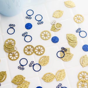 May include: A scattering of gold glitter lemon slices, leaves, and blue glitter engagement ring cutouts on a white surface. Two light blue cups are in the background. The items are likely for a wedding or party.