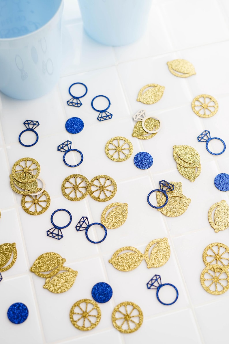 May include: A scattering of gold glitter lemon slices, leaves, and blue glitter engagement ring cutouts on a white surface. Two light blue cups are in the background. The items are likely for a wedding or party.