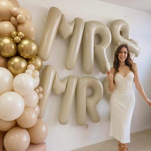 Könnte beinhalten: Eine Braut posiert vor beigen "MRS"-Ballons. Sie trägt ein weißes Kleid und hält ein Sektglas. Links befindet sich ein Ballonbogen in Beige- und Goldtönen.