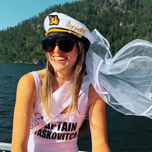 Puede incluir: Una novia con un sombrero de capit&aacute;n blanco con la inscripci&oacute;n "bride" en dorado y un velo blanco. Tambi&eacute;n lleva una camiseta blanca de tirantes con el texto "Captain Askowitch" impreso.
