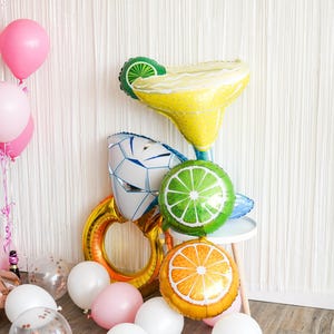 May include: A festive arrangement of foil balloons in the shape of a margarita, a lime slice, an orange slice, and a diamond ring. Pink, white, and clear balloons with confetti are scattered on the floor, creating a celebratory atmosphere.