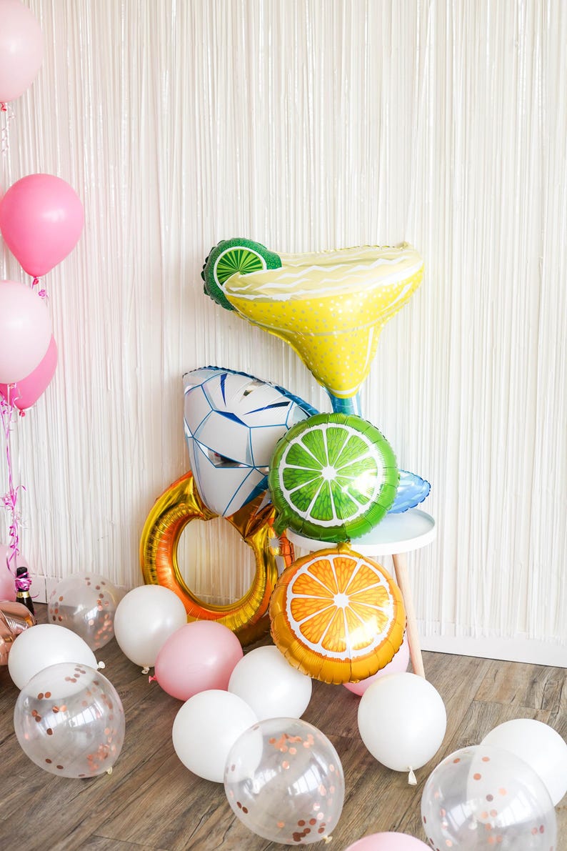 May include: A festive arrangement of foil balloons in the shape of a margarita, a lime slice, an orange slice, and a diamond ring. Pink, white, and clear balloons with confetti are scattered on the floor, creating a celebratory atmosphere.