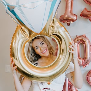 May include: A large diamond ring balloon with a gold band and a white and blue diamond shape. The balloon is held up in front of a person. Pink and gold balloons spell out "I DO" on the wall.