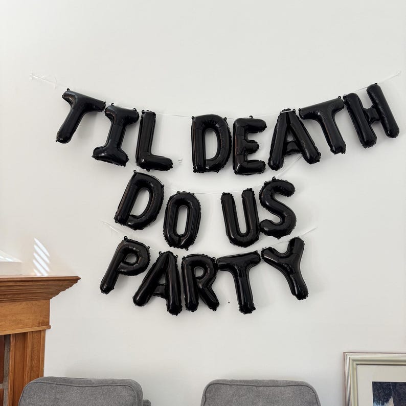 May include: A set of black foil letter balloons arranged in three rows on a wall, spelling out the phrase TIL DEATH DO US PARTY. The balloons are inflated and connected by a thin white string, creating a bold, dark decorative display.