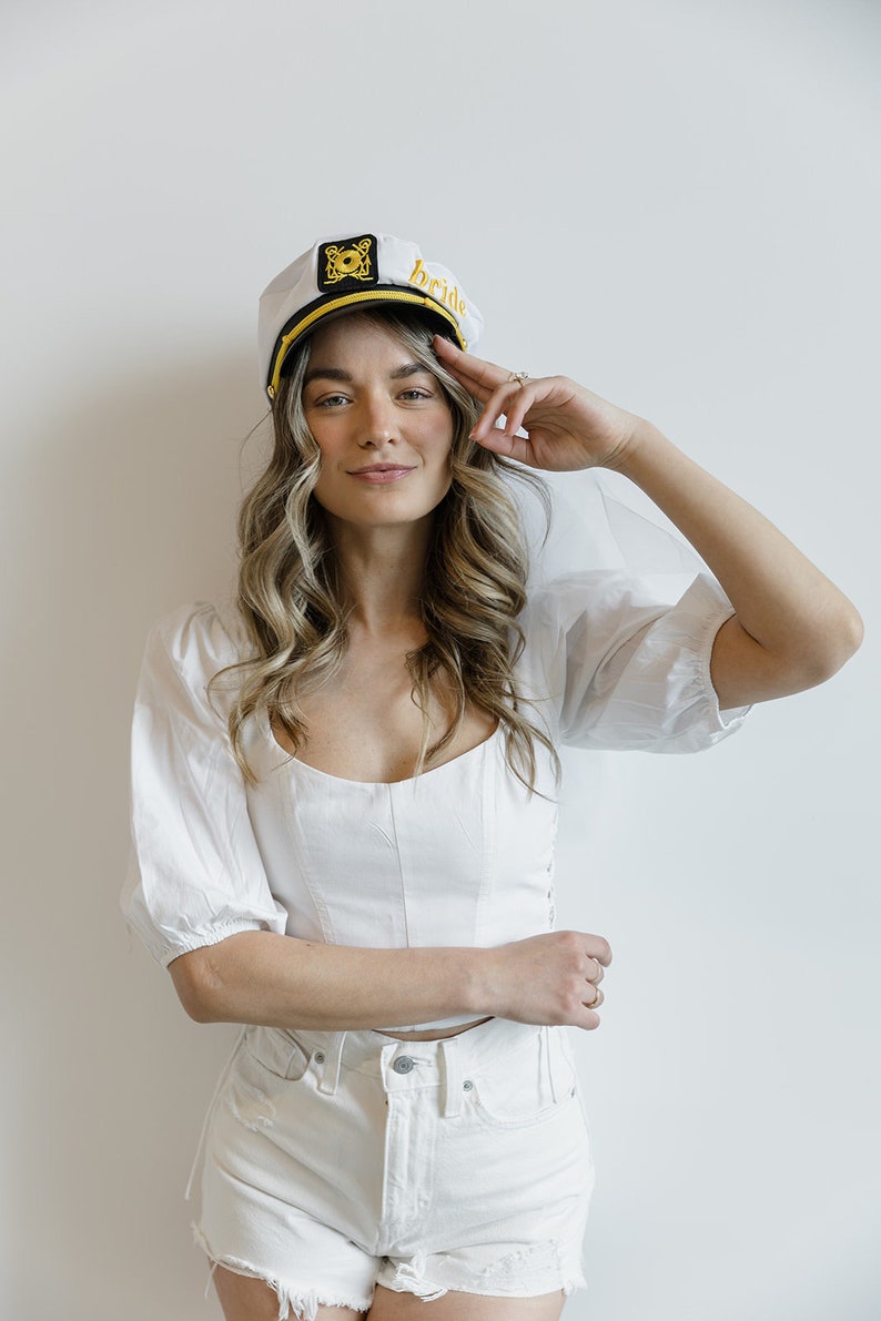 Puede incluir: Una mujer con un sombrero de marinero blanco con la palabra "bride" bordada, una blusa blanca y pantalones cortos de mezclilla blancos. Est&aacute; sonriendo y saludando con la mano derecha.