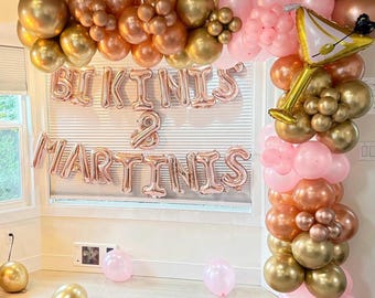 May include: A pink and gold balloon arch with the words "Bikinis & Martinis" in gold foil letters. The arch is decorated with a gold balloon shaped like a martini glass.