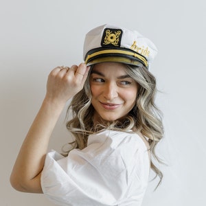 Puede incluir: Un sombrero de marinero blanco con ribete dorado y la palabra "bride" bordada en un lateral. El sombrero tiene un ancla negra y un dise&ntilde;o circular dorado en la parte delantera.