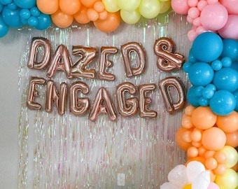 May include: A backdrop of iridescent foil fringe with a balloon arch in shades of blue, yellow, orange, and pink. The balloon arch is decorated with a large rose gold foil sign that reads "DAZED & ENGAGED".