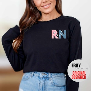 May include: A black long-sleeved shirt with the letters "RN" in pink and blue with the word "Kylie" written in white script inside the letters. The shirt is being worn by a person with long brown hair. The text "FRAY COLLECTIVE ORIGINAL DESIGNER" is visible in the lower right corner of the image.
