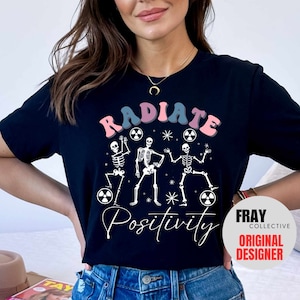 May include: Black t-shirt with a white graphic of three skeletons dancing and holding radioactive symbols. The text "RADIATE POSITIVITY" is printed in pink, blue, and white. The Fray Collective Original Designer logo is in the bottom right corner.