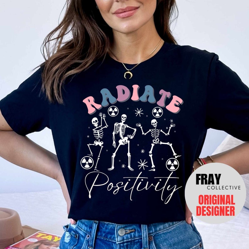 May include: Black t-shirt with a white graphic of three skeletons dancing and holding radioactive symbols. The text "RADIATE POSITIVITY" is printed in pink, blue, and white. The Fray Collective Original Designer logo is in the bottom right corner.