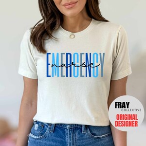 Puede incluir: Una camiseta blanca con el texto "EMERGENCY nurse" en letras azules y negras. El texto est&aacute; apilado verticalmente con la palabra "nurse" escrita en cursiva.