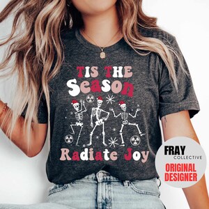 May include: A gray t-shirt with the text "Tis the Season to Radiate Joy" in pink and red letters. Three skeletons wearing Santa hats are dancing around a radioactive symbol. The text "Fray Collective Original Designer" is in a white circle with a red border.