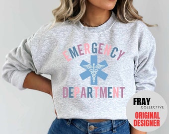 May include: A gray sweatshirt with the text "EMERGENCY DEPARTMENT" in pink, blue, and gray lettering. A blue medical symbol with a white snake and wings is centered between the words. The text "FRAY COLLECTIVE ORIGINAL DESIGNER" is in a white circle with a red border.
