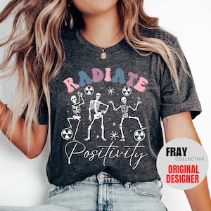 May include: A gray t-shirt with a graphic of three skeletons dancing with the text "RADIATE POSITIVITY" in pink, blue, and white. The graphic also includes several white stars and pink and blue radiation symbols. The text "FRAY COLLECTIVE ORIGINAL DESIGNER" is in a white circle with a red border.