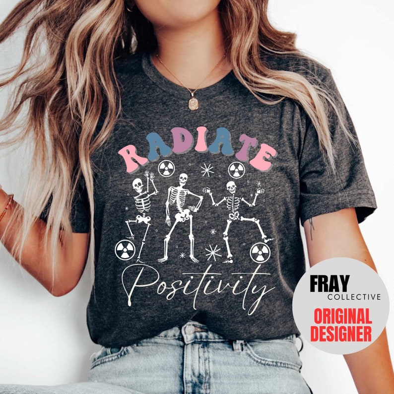 May include: A gray t-shirt with a graphic of three skeletons dancing with the text "RADIATE POSITIVITY" in pink, blue, and white. The graphic also includes several white stars and pink and blue radiation symbols. The text "FRAY COLLECTIVE ORIGINAL DESIGNER" is in a white circle with a red border.