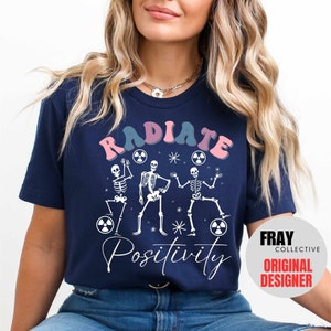 May include: A navy blue t-shirt with a graphic of three skeletons dancing with the text "RADIATE POSITIVITY" in pink, blue, and white. The graphic also includes a white star and three white radiation symbols. The t-shirt has a Fray Collective Original Designer label.