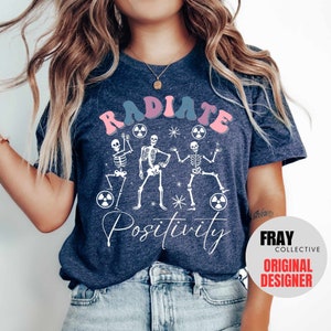 May include: A navy blue t-shirt with a white graphic of three skeletons dancing with the text "RADIATE POSITIVITY" in pink and blue letters.