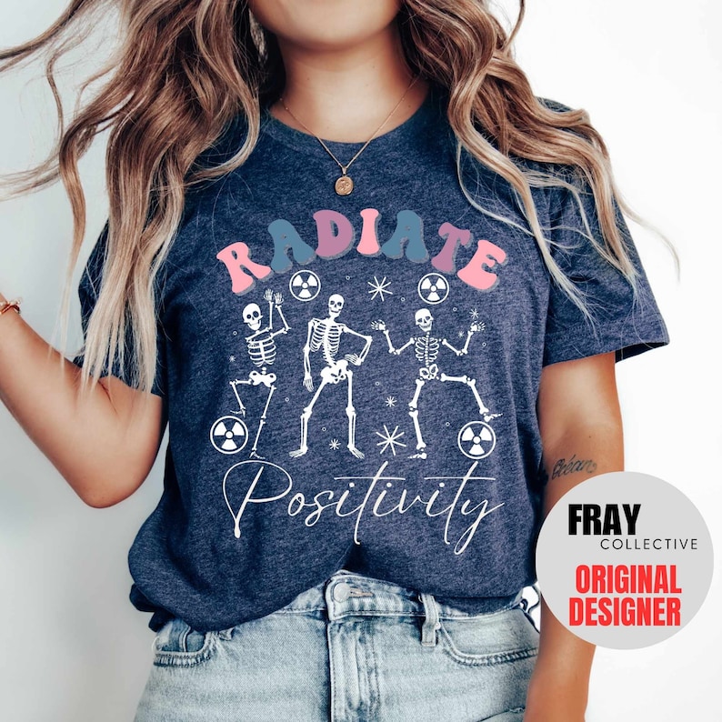 May include: A navy blue t-shirt with a white graphic of three skeletons dancing with the text "RADIATE POSITIVITY" in pink and blue letters.