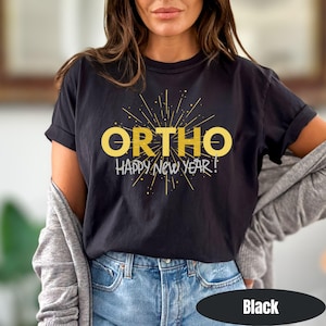 May include: Black t-shirt with the word "ORTHO" in gold letters, and the phrase "HAPPY NEW YEAR!" in white. The design features a starburst pattern. The shirt is paired with blue jeans.