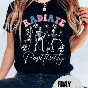 May include: Black t-shirt with a graphic of three skeletons dancing with the text "RADIATE POSITIVITY" in pink, blue, and purple letters. The skeletons are holding radiation symbols.