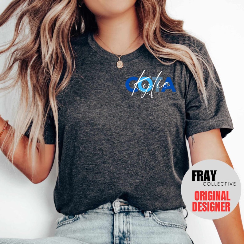 May include: A dark gray t-shirt with a blue and white graphic design that says "Kylie" on the front. The shirt is being worn by a person with long blonde hair. The image also includes a logo for "Fray Collective Original Designer".