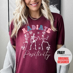 May include: A maroon t-shirt with a white graphic of three skeletons dancing with the text "RADIATE POSITIVITY" in pink, blue, and white. The text "FRAY COLLECTIVE ORIGINAL DESIGNER" is in a white circle on the bottom right corner of the shirt.