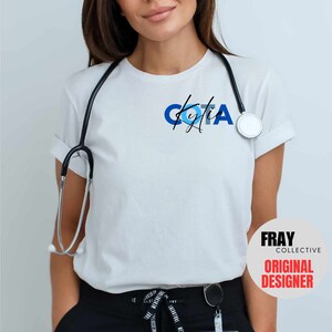 May include: White t-shirt with a blue and black design that says "COTA" with a signature underneath. The shirt is worn by a person with a stethoscope around their neck. The text "FRAY COLLECTIVE ORIGINAL DESIGNER" is in a white circle with a black outline.