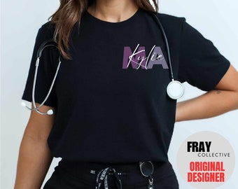 Puede incluir: Camiseta negra con un gráfico morado y blanco que dice "MA" con el nombre "Kylie" escrito en cursiva arriba. La camiseta la lleva puesta una persona con un estetoscopio alrededor del cuello. El texto "FRAY COLLECTIVE ORIGINAL DESIGNER" es visible en la esquina inferior derecha de la imagen.
