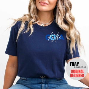 May include: A navy blue t-shirt with a white graphic of the letters "OTA" in blue and white circles. The name "Kylie" is written in white script below the letters. The text "FRAY COLLECTIVE ORIGINAL DESIGNER" is visible in the bottom right corner of the image.