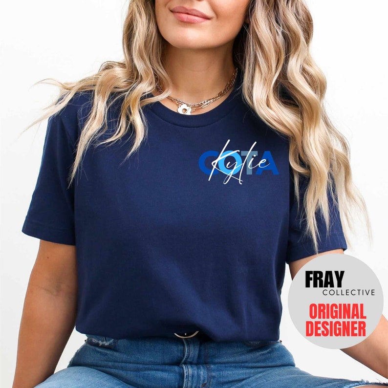May include: A navy blue t-shirt with a white graphic of the letters "OTA" in blue and white circles. The name "Kylie" is written in white script below the letters. The text "FRAY COLLECTIVE ORIGINAL DESIGNER" is visible in the bottom right corner of the image.