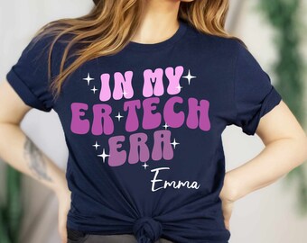 May include: Navy blue t-shirt with the text "In My ER Tech Era" in pink and white with glitter accents. The name "Emma" is written in white below the text.