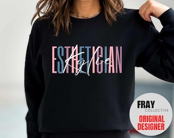 May include: Black sweatshirt with the text "ESTHETICIAN" in pink, blue, and gray vertical lettering. The word "Kylie" is written in cursive in white over the text. The text "FRAY COLLECTIVE ORIGINAL DESIGNER" is in a white circle on the bottom right corner of the sweatshirt.