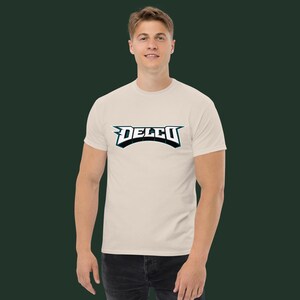 Peut inclure: T-shirt beige avec le mot "DELCO" en police stylisée sur la poitrine. Le texte est souligné en noir et bleu sarcelle. Le t-shirt est à manches courtes et porté par une personne.