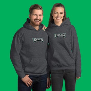 Puede incluir: Dos personas vistiendo sudaderas con capucha de color gris oscuro con la palabra "DELCO" impresa en verde en la parte delantera. Las sudaderas tienen una capucha con cordón y un bolsillo canguro.