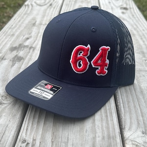 May include: A navy blue trucker hat with a red and white embroidered number "64" on the front. The hat has a black mesh back and a black adjustable strap.