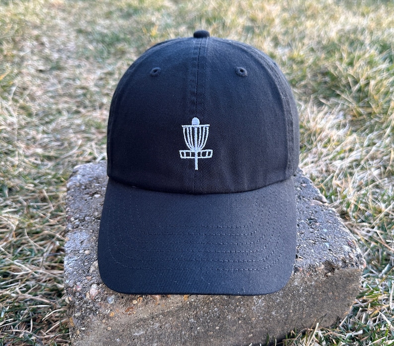K&ouml;nnte beinhalten: Eine schwarze Baseballkappe mit einem wei&szlig;en gestickten Disc-Golf-Korb-Design.