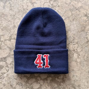 May include: A navy blue knit beanie with a folded brim. The number "41" is embroidered in red with a white outline on the front of the folded brim. The beanie is made of a soft, knitted material.