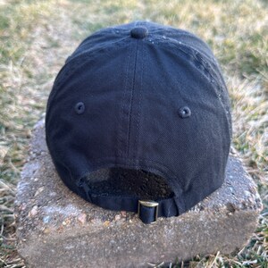 K&ouml;nnte beinhalten: Schwarze Baseballkappe mit einem schwarzen verstellbaren Riemen und zwei kleinen schwarzen &Ouml;sen auf der R&uuml;ckseite.
